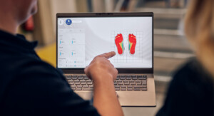 Ein Laptopbildschirm zeigt farbige Druckverteilungen beider Füße, eine Person deutet mit dem Finger darauf, während eine zweite Person zusieht.