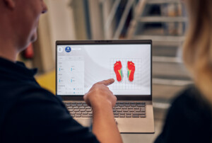Ein Laptopbildschirm zeigt farbige Druckverteilungen beider Füße, eine Person deutet mit dem Finger darauf, während eine zweite Person zusieht.
