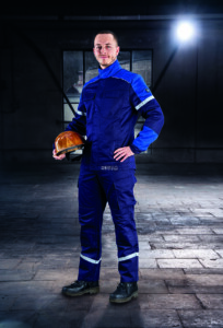 Ein Mann in dunkelblauer Schutzkleidung mit Reflexstreifen hält einen gelben Schutzhelm in der Hand und steht in einer schwach beleuchteten Industriehalle.