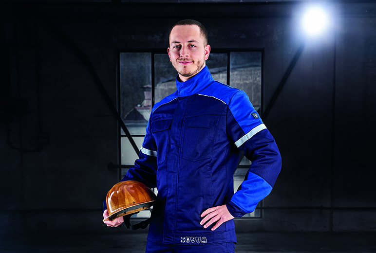 Ein Mann in dunkelblauer Schutzkleidung mit Reflexstreifen hält einen gelben Schutzhelm in der Hand und steht in einer schwach beleuchteten Industriehalle.