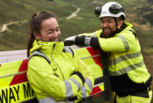 Eine Arbeiterin und ein Arbeiter in gelben Warnjacken lehnen lachend an ein Arbeitsauto vor grüner Landschaft. Der Mann trägt einen weißen Schutzhelm.
