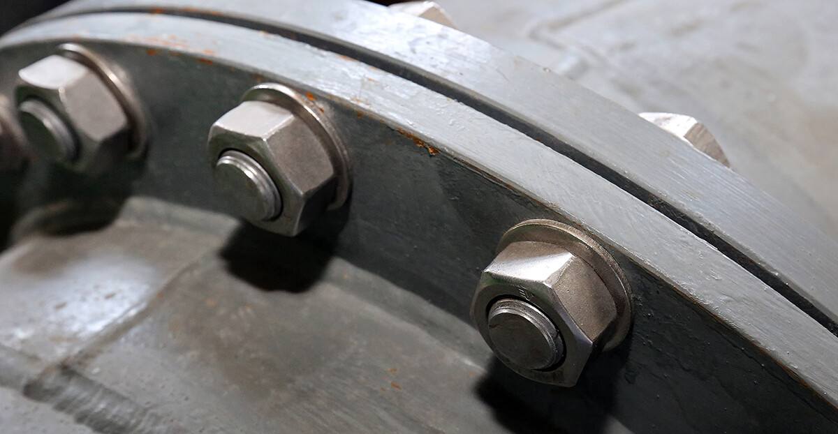 Ein Detailfoto zeigt eine Rohrflanschverbindung mit mehreren großen Sechskantschrauben und Muttern auf grauer Metalloberfläche.
