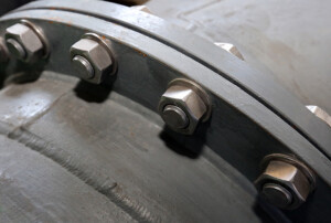 Ein Detailfoto zeigt eine Rohrflanschverbindung mit mehreren großen Sechskantschrauben und Muttern auf grauer Metalloberfläche.