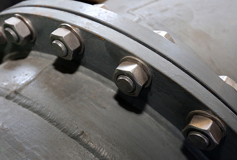 Ein Detailfoto zeigt eine Rohrflanschverbindung mit mehreren großen Sechskantschrauben und Muttern auf grauer Metalloberfläche.
