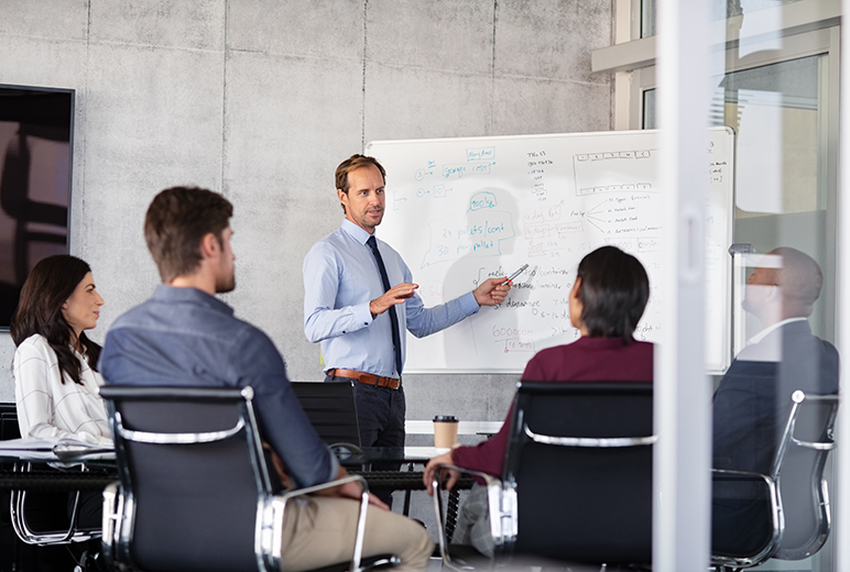 Ein Mann präsentiert vor einem Whiteboard mit Diagrammen und handschriftlichen Notizen, während vier Kolleginnen und Kollegen zuhören.