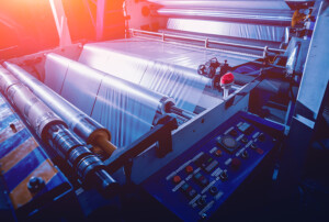 Eine Folienproduktionsanlage zeigt breite Kunststofffolie, die über mehrere Walzen läuft, daneben befindet sich ein Steuerpult mit Bedienelementen und rotem Not-Aus-Taster.