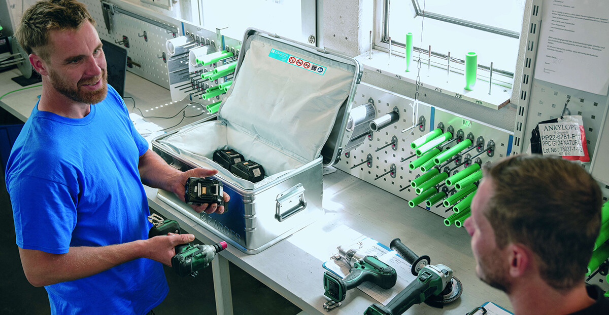 In einer Werkstatt hält ein Mitarbeiter im blauen Shirt einen Akkupack hoch, auf der Werkbank stehen ein offener Aluminiumkoffer mit weiteren Lithium-Batterien und mehrere Elektrowerkzeuge.