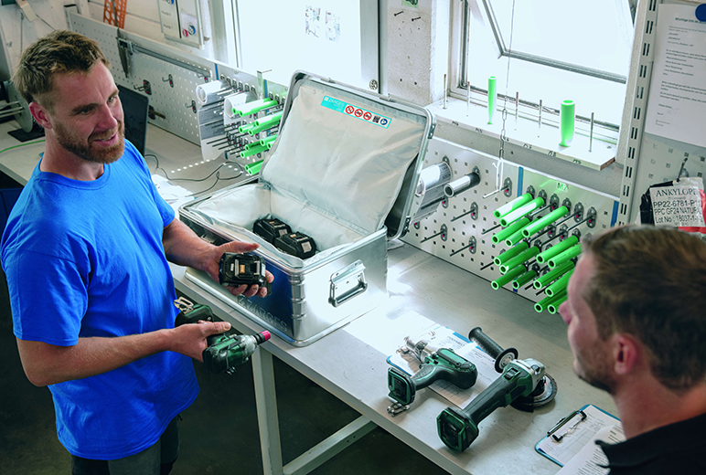 In einer Werkstatt hält ein Mitarbeiter im blauen Shirt einen Akkupack hoch, auf der Werkbank stehen ein offener Aluminiumkoffer mit weiteren Lithium-Batterien und mehrere Elektrowerkzeuge.