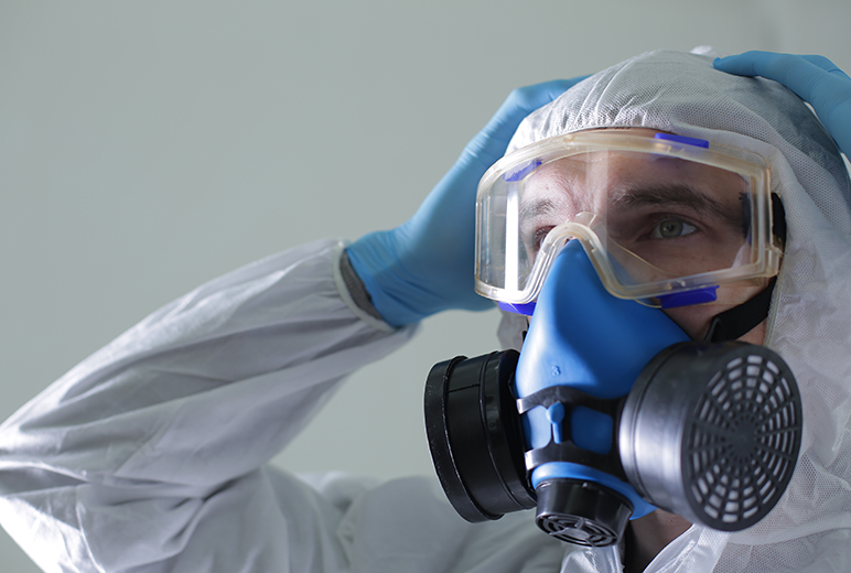 Eine Person in Schutzanzug und blauen Einweghandschuhen trägt eine Vollgesichtsatemschutzmaske mit Filtern und hält beide Hände an den Kopf.