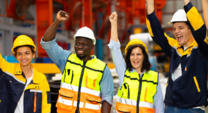 Vier Beschäftigte in Helmen und Warnwesten jubeln gemeinsam in einer Werkhalle.