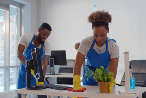 Zwei Reinigungskräfte mit Handschuhen säubern Schreibtische in einem hellen Büro, Monitore und Putzmittel sind zu sehen.