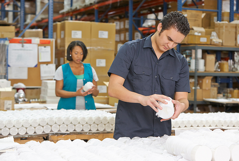 Zwei Beschäftigte prüfen verpackte Produkte an einem Arbeitstisch im Warenlager; im Hintergrund stehen hohe Regale und gestapelte Kartons.