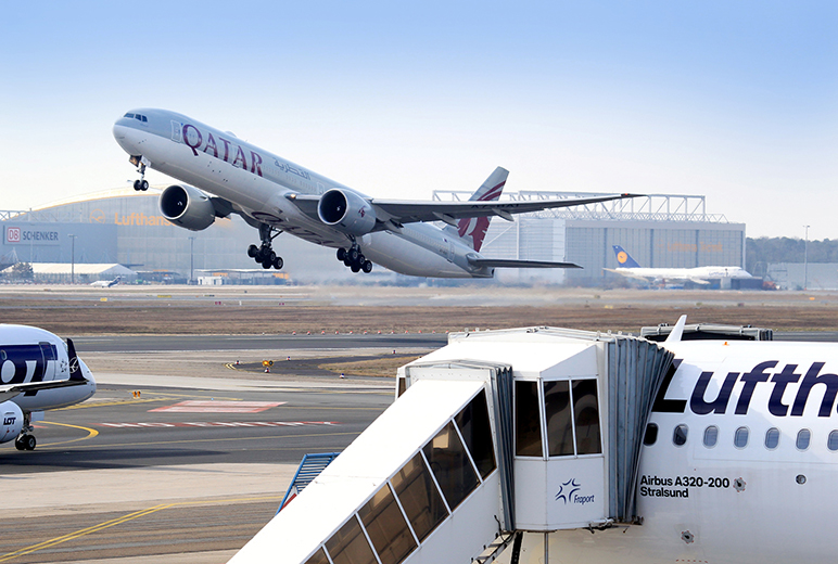 Am Flughafen hebt ein Qatar-Airways-Jet ab, im Vordergrund steht ein Lufthansa-Flugzeug mit angedockter Fluggastbrücke.