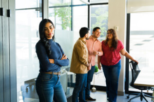Eine Frau steht allein in einem Büro, während im Hintergrund drei Personen miteinander sprechen und lachen.