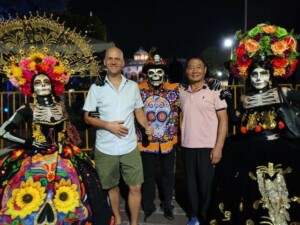 Bei Nacht posieren zwei prächtig verkleidete Catrinas und ein Skelett zwischen zwei Besuchern auf einem belebten Platz.