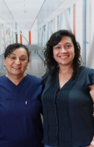 Drei Personen im Klinikflur, zwei in dunkelblauen Pflegeuniformen und eine in ziviler Kleidung, lächeln nebeneinander in die Kamera.